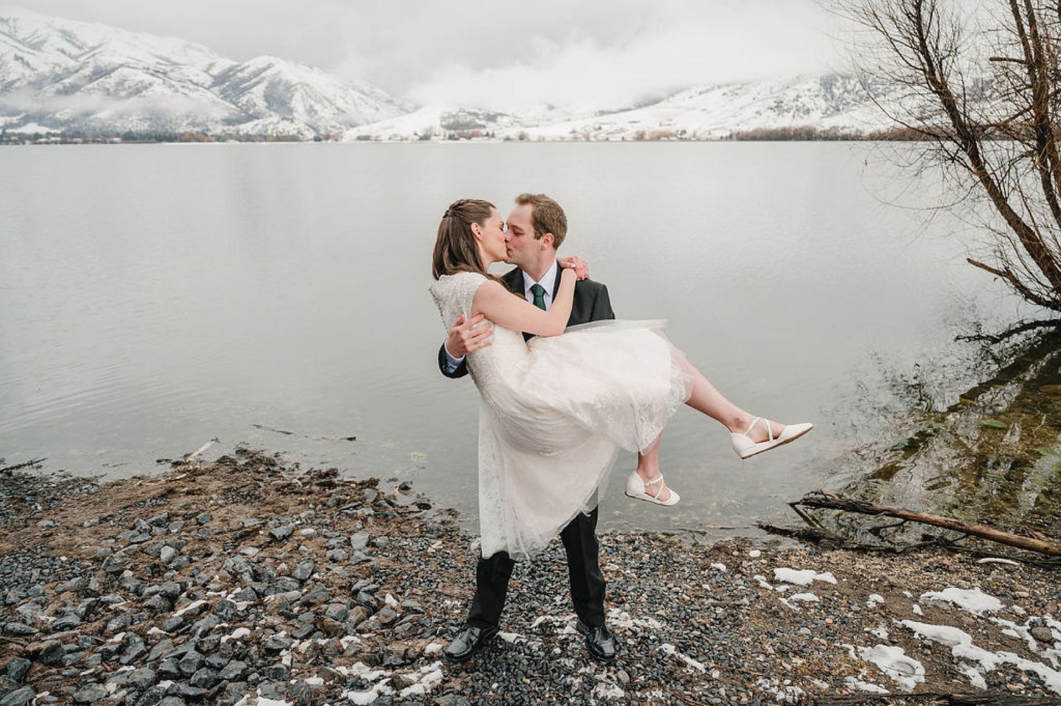 A couple shares a romantic moment by a snowy mountain lake as the groom carries the bride in her flowing white dress.