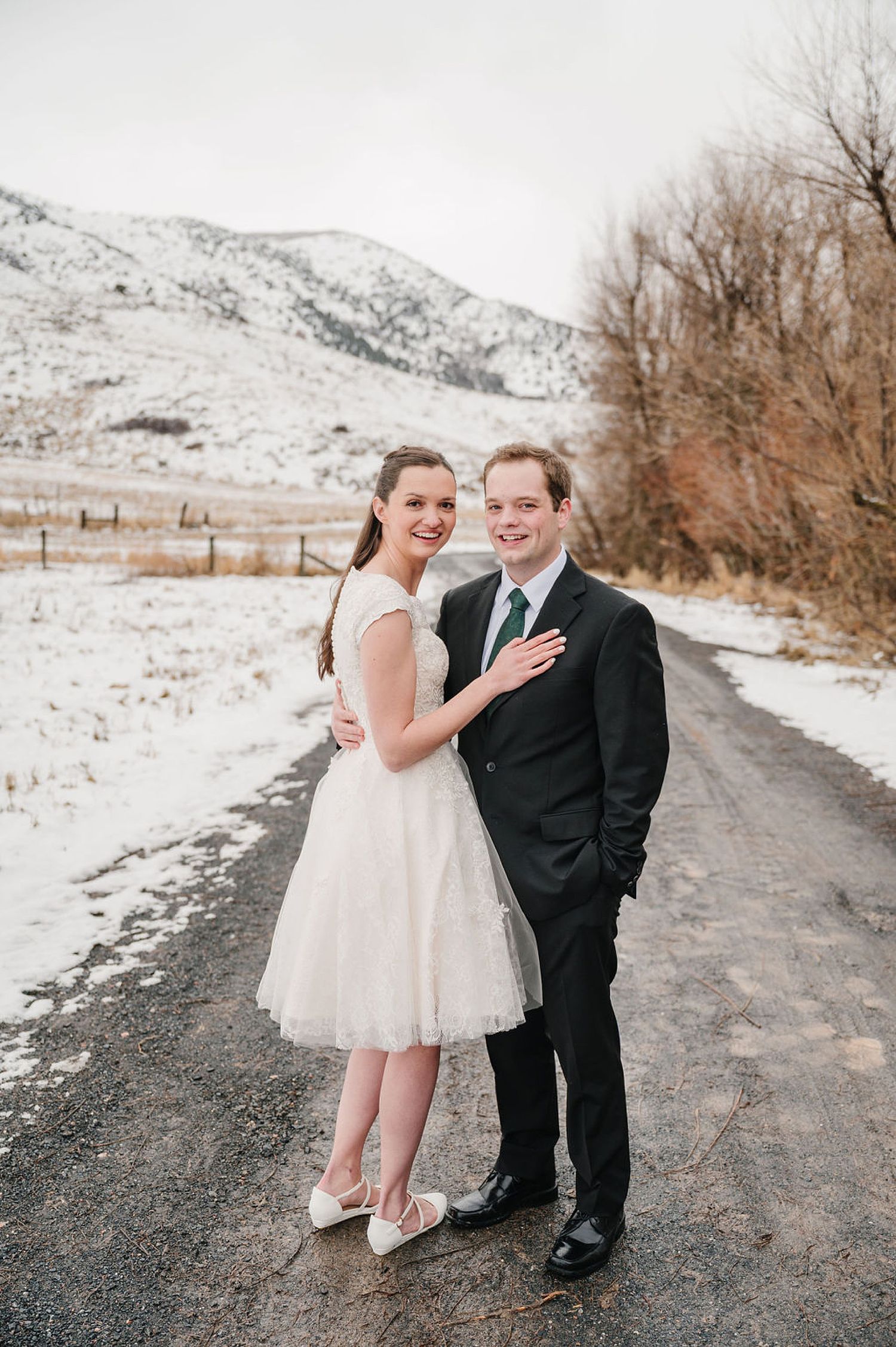 A couple poses together on a snowy mountain road, the bride wearing a knee-length white dress and the groom in a black suit.