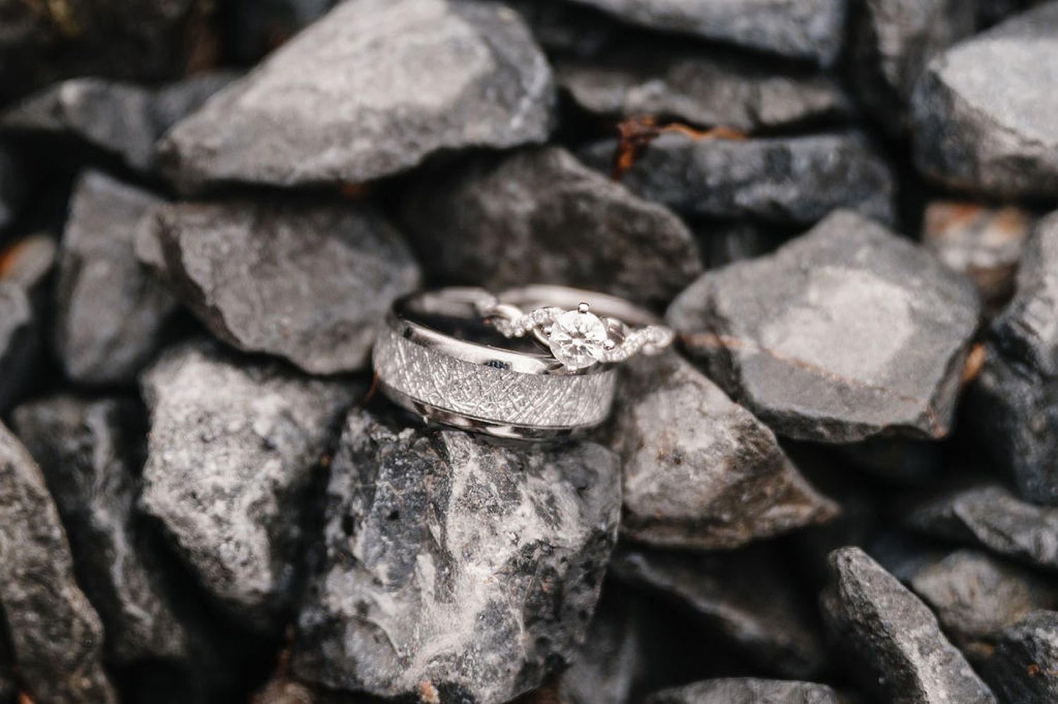 Two elegant wedding rings with diamond details rest on a bed of gray stones in dramatic lighting.