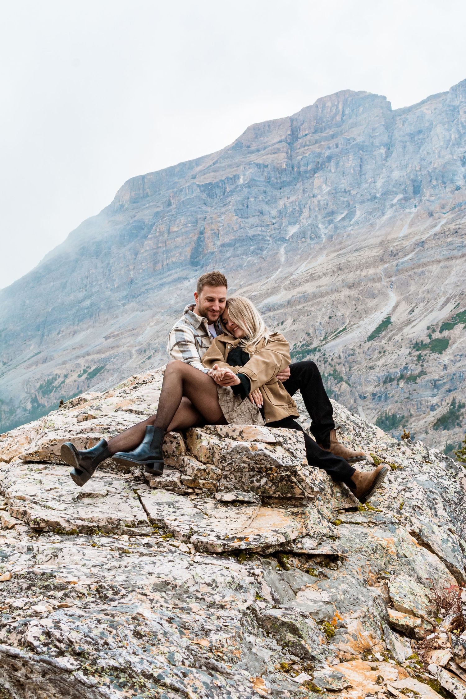 A couple shares a romantic moment on a mountain overlook with stunning rocky peaks and forest valleys in the background.