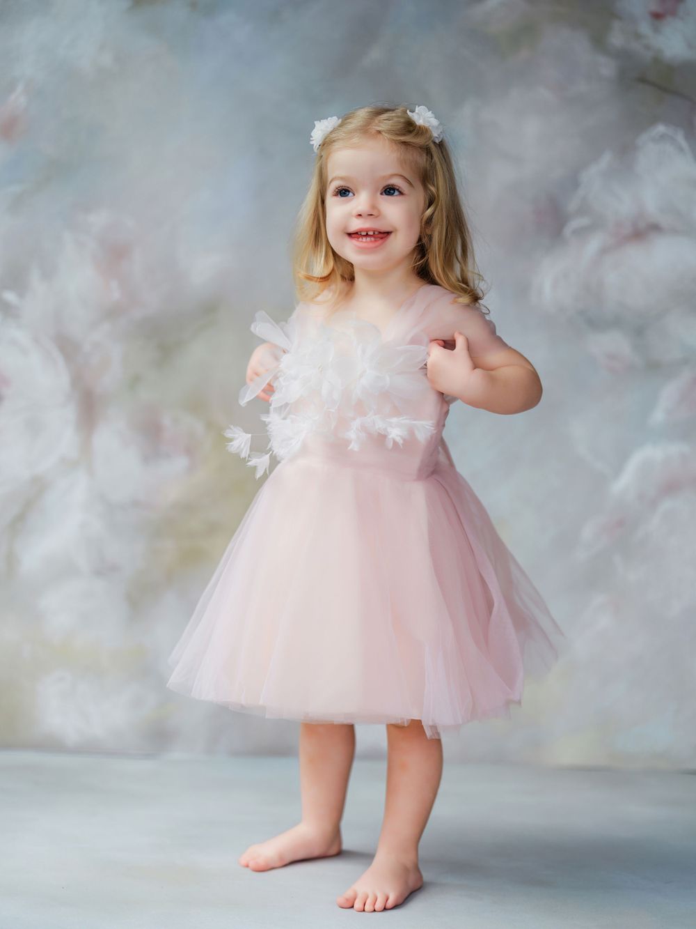 Small blonde child wearing a light pink tulle dress with lace details poses barefoot against a soft cloudy backdrop in Bellevue studio.