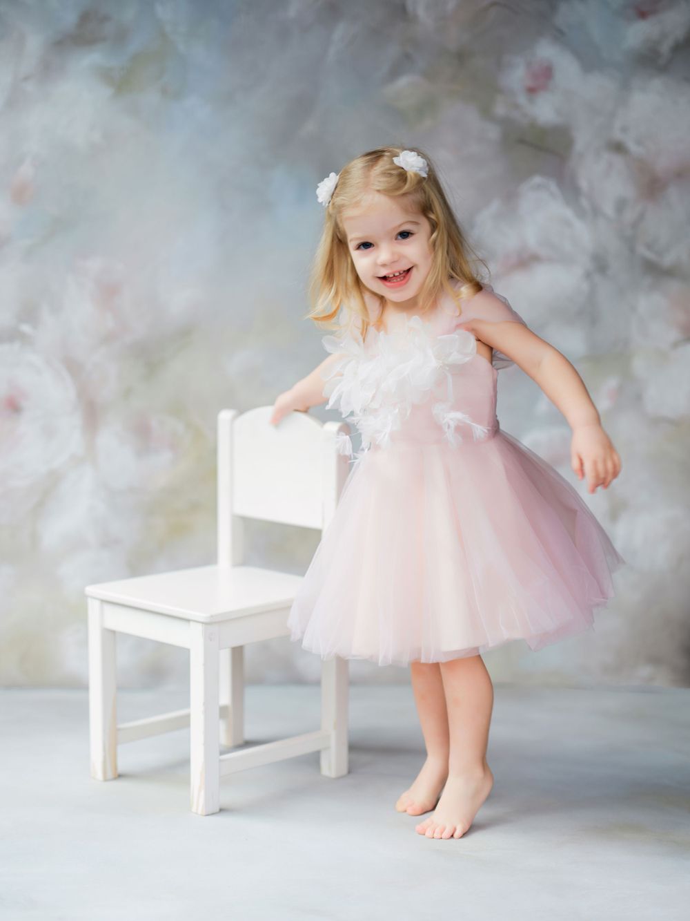 A small child in a pink tulle dress dances barefoot next to a white chair against a dreamy painted backdrop in Bellevue studio.