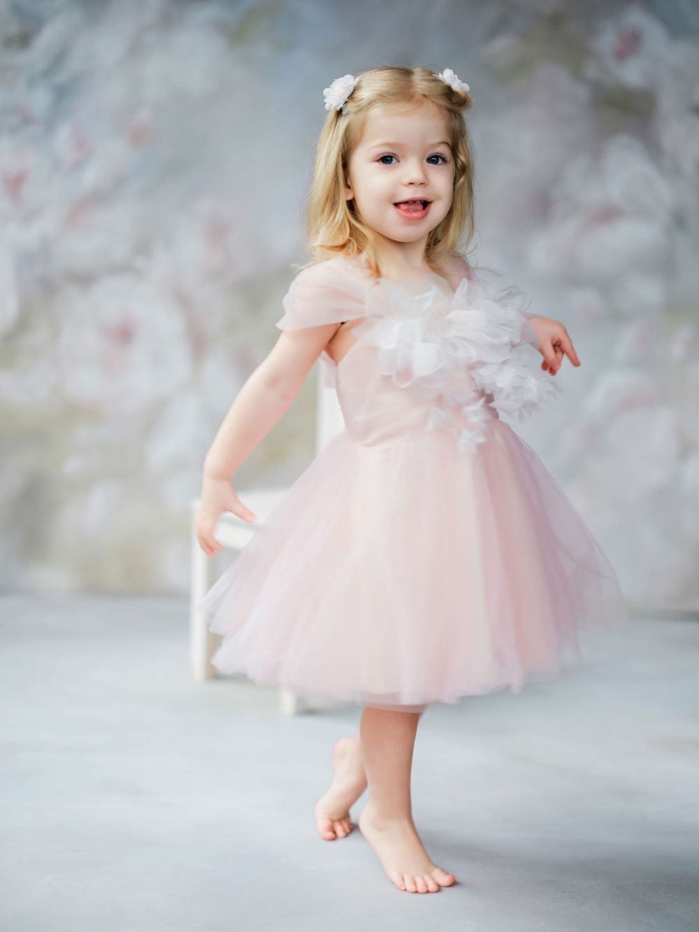 A young dancer in a ballerina-style pink tulle dress and white hair bows poses gracefully on a soft gray background in Bellevue studio.