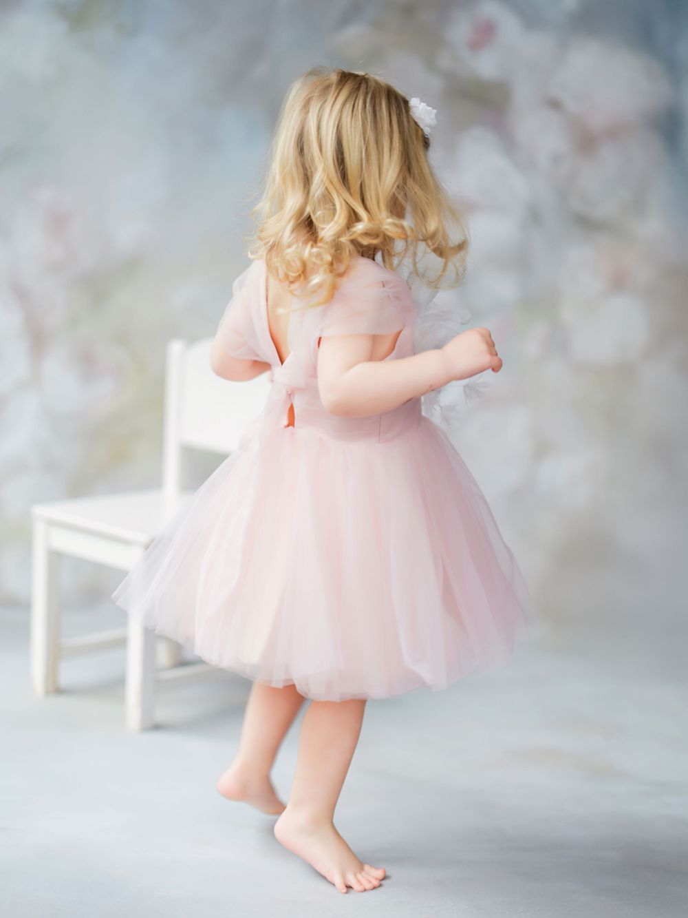 Little girl in a pink tulle dress twirls barefoot against a dreamy gray backdrop with a white chair in Bellevue studio.