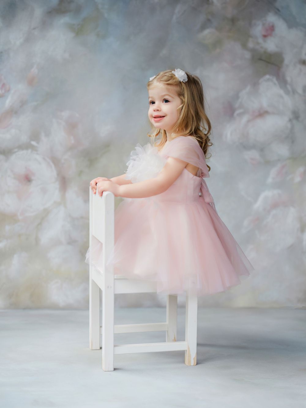 A child in a soft pink party dress stands beside a white wooden chair against a mottled gray background in Bellevue studio.