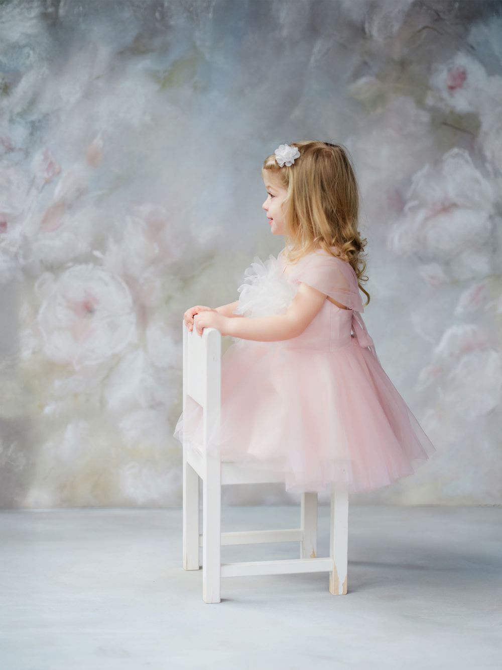 Small child in pink tulle dress stands on white chair against dreamy painted background in Bellevue studio.