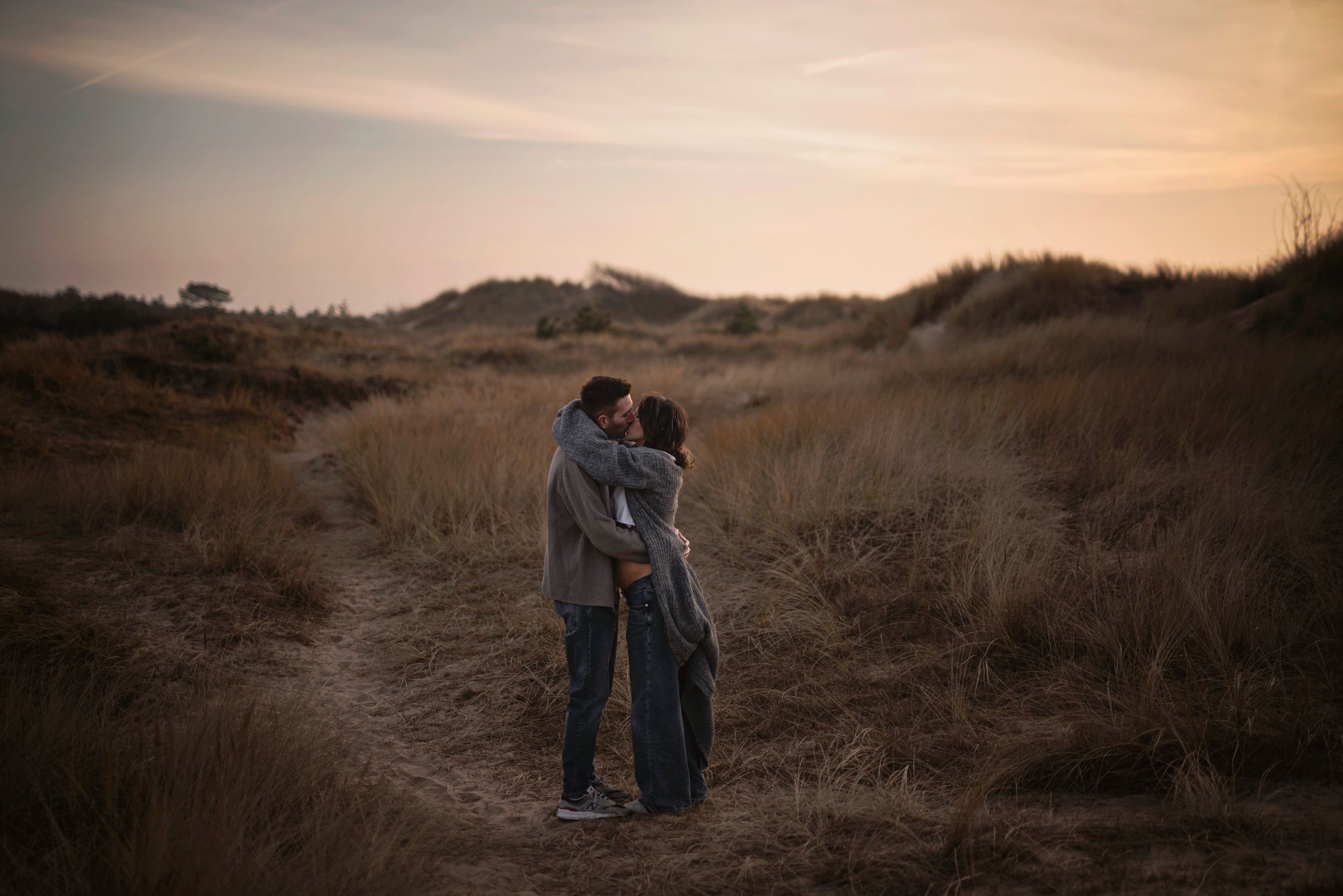 A couple embraces in a golden field at sunset with tall grasses and rolling hills in the background.