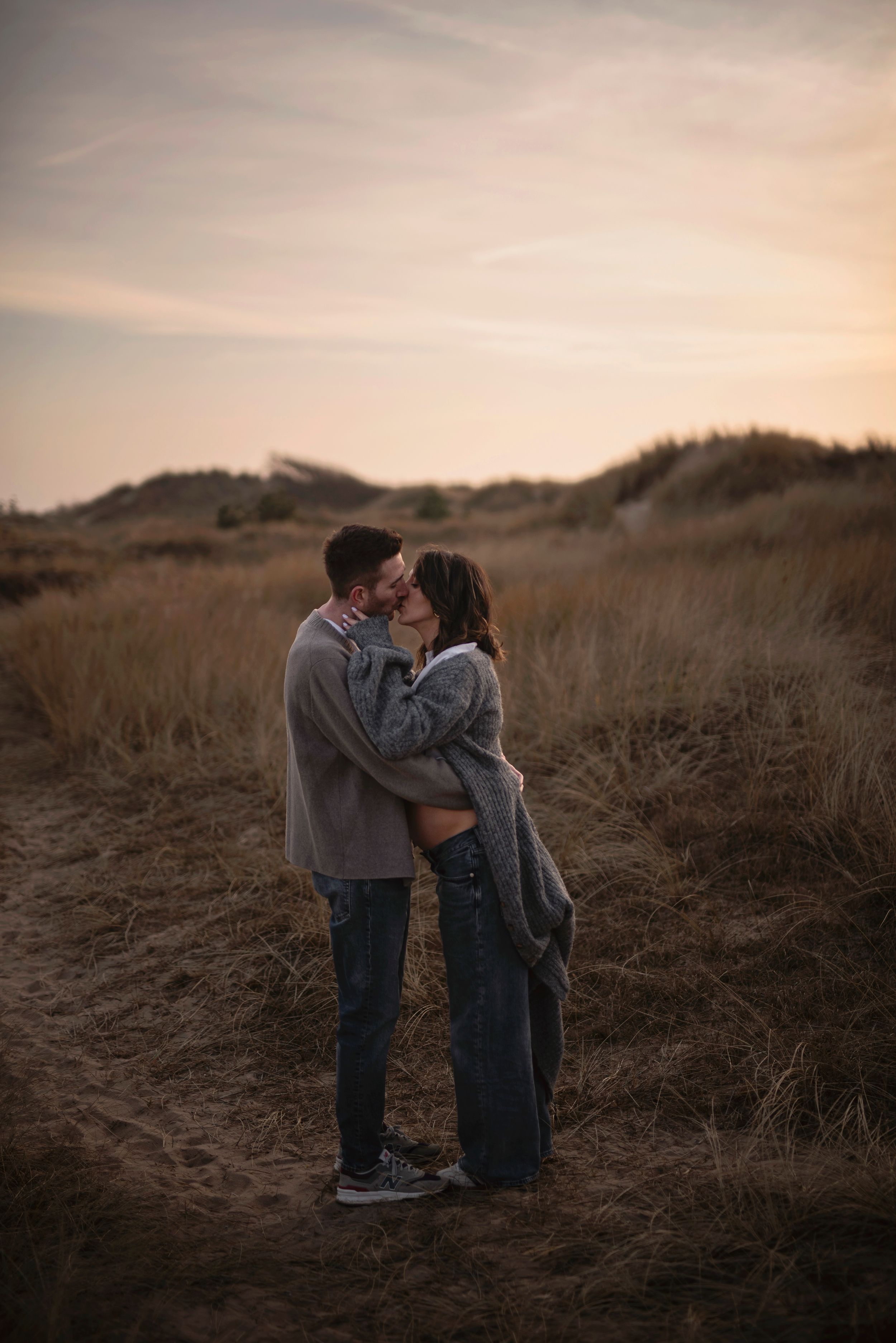 A couple shares a romantic moment at sunset among tall grasses on a hillside path.