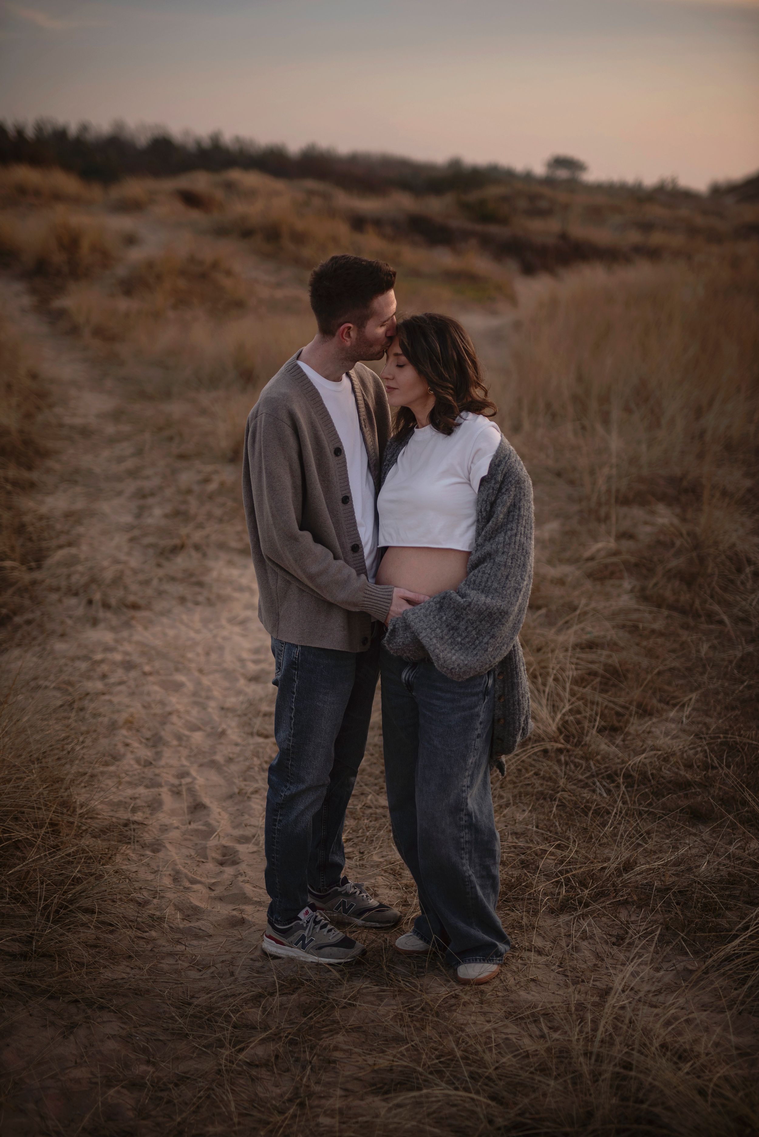 Couple embraces on a dirt path during golden hour in a rustic outdoor setting with dry brown grass and hills.
