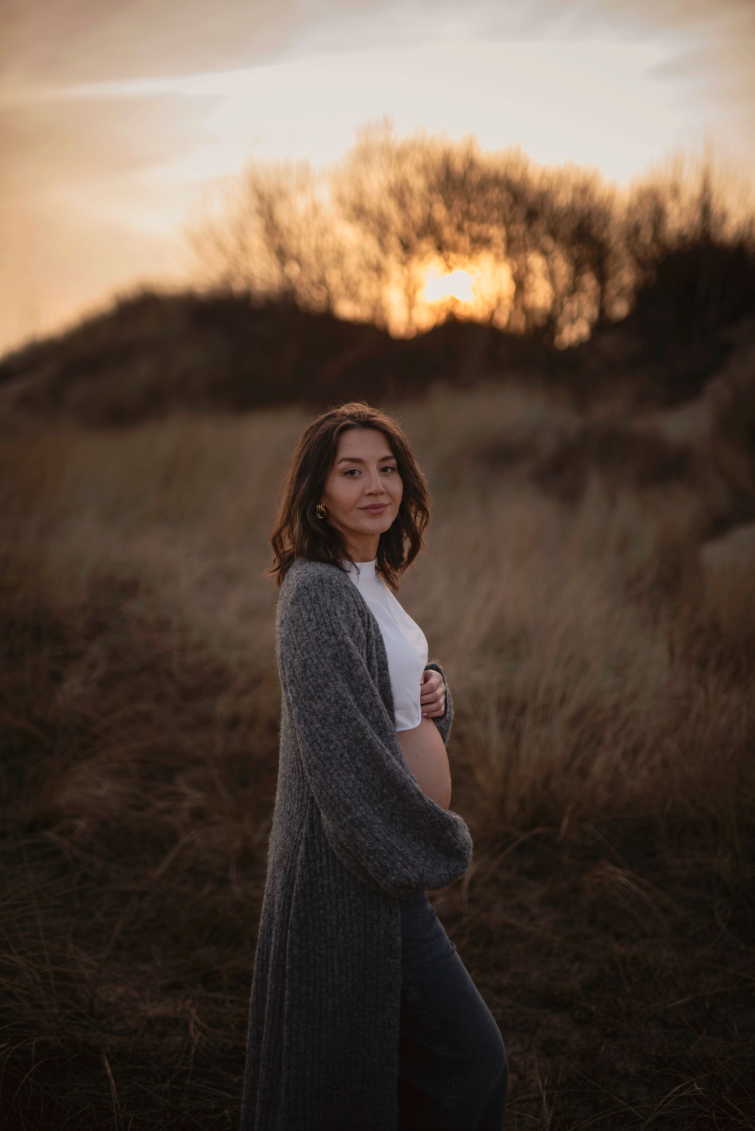 Expectant mother in gray cardigan stands against sunset with silhouetted trees in background at golden hour.