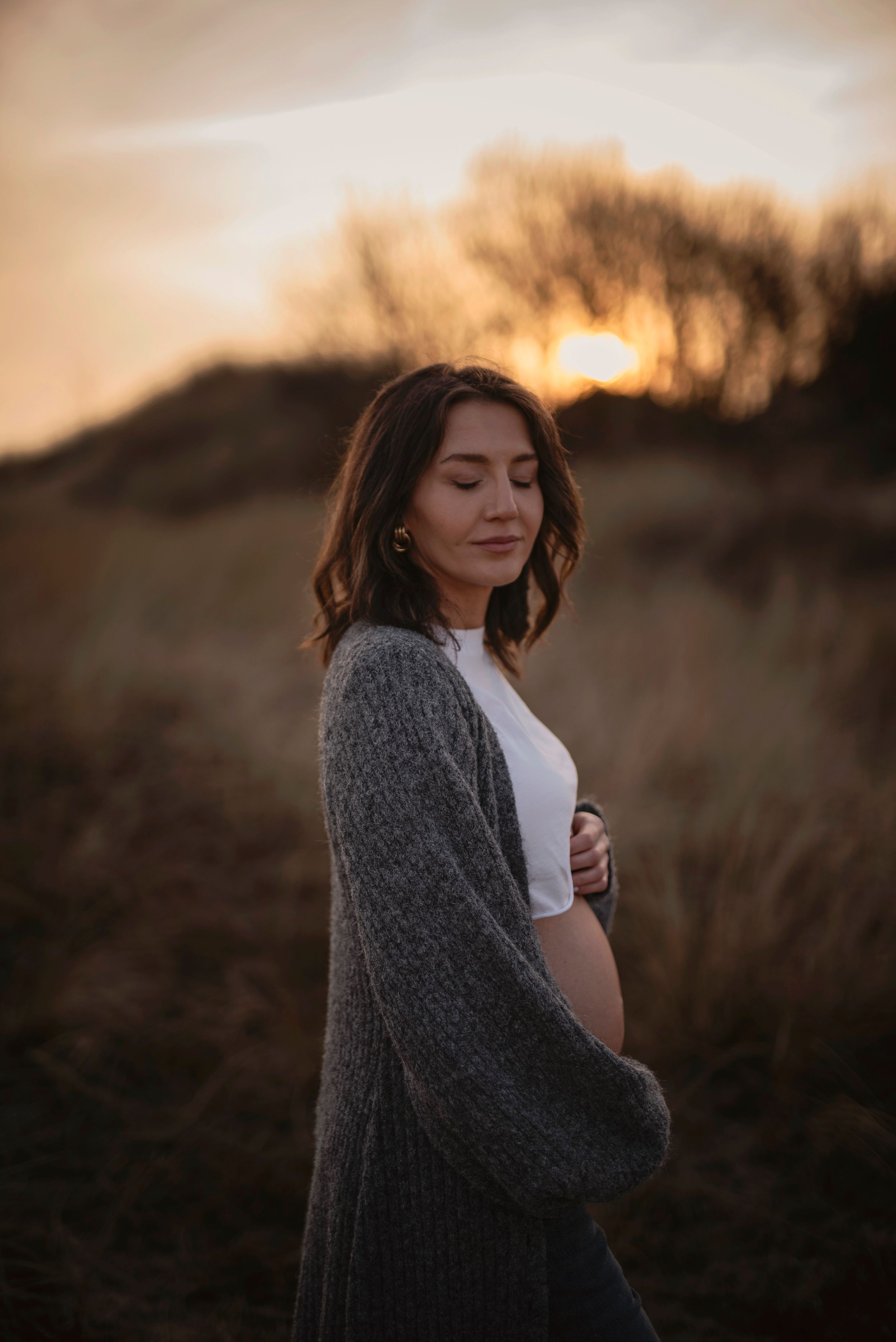 A contemplative person in a gray cardigan stands in a field at sunset with warm golden light behind them.