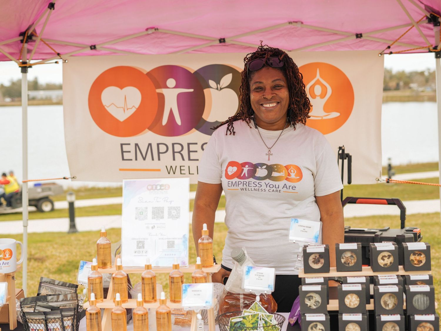 A vendor stands behind a display of products at an outdoor market under a pink tent with wellness company branding.