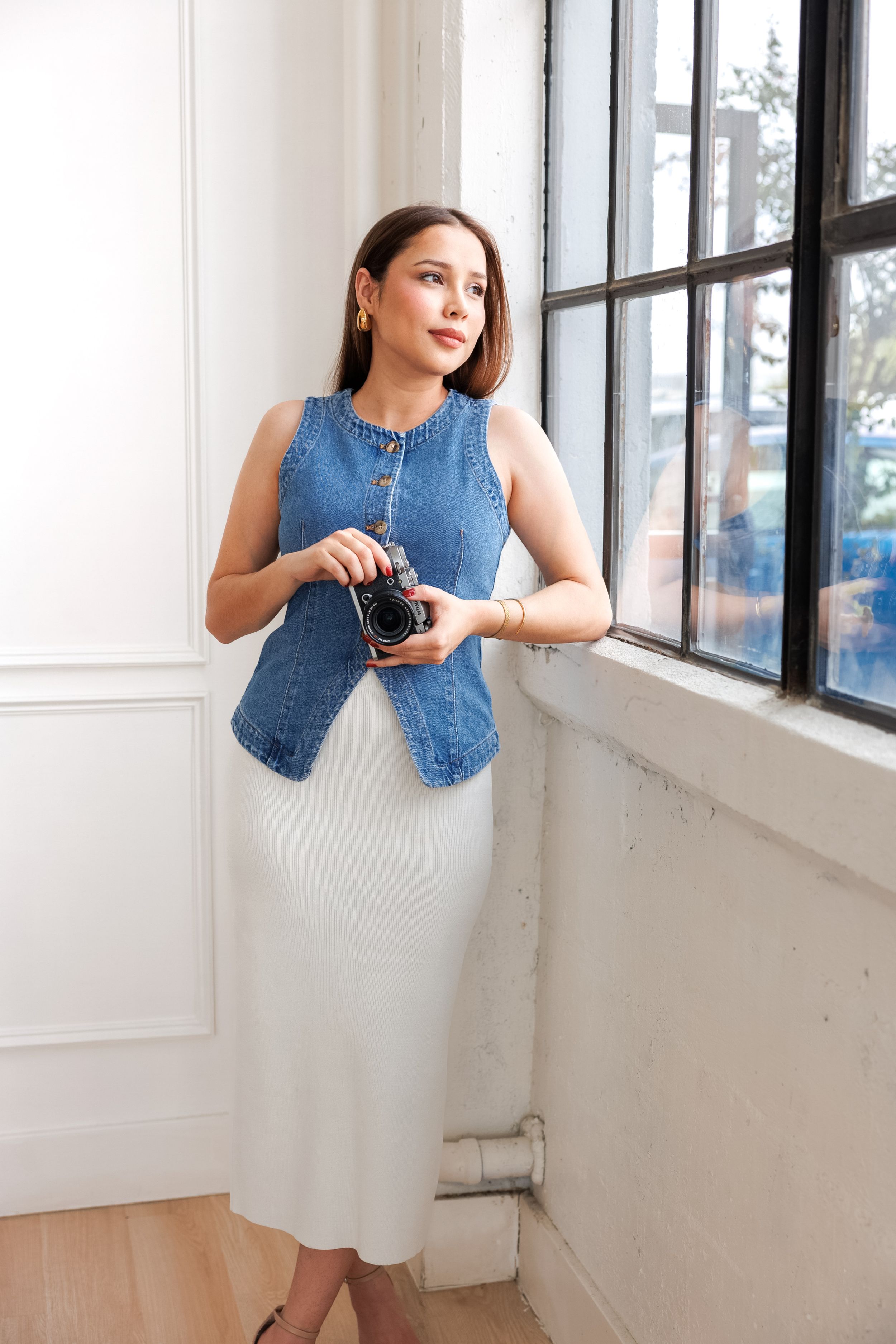 Photographer in blue and white striped button-down shirt holding vintage camera by window in casual indoor setting.