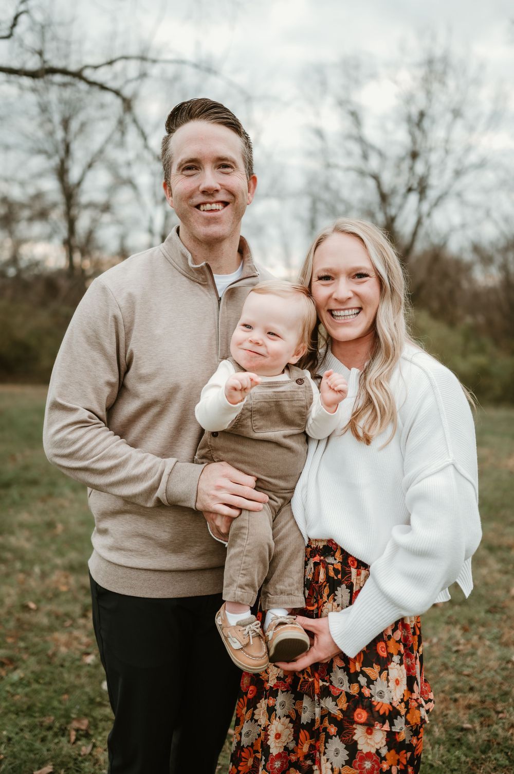 Happy young family with a baby boy posing outdoors in autumn, wearing cozy sweaters and a floral skirt.