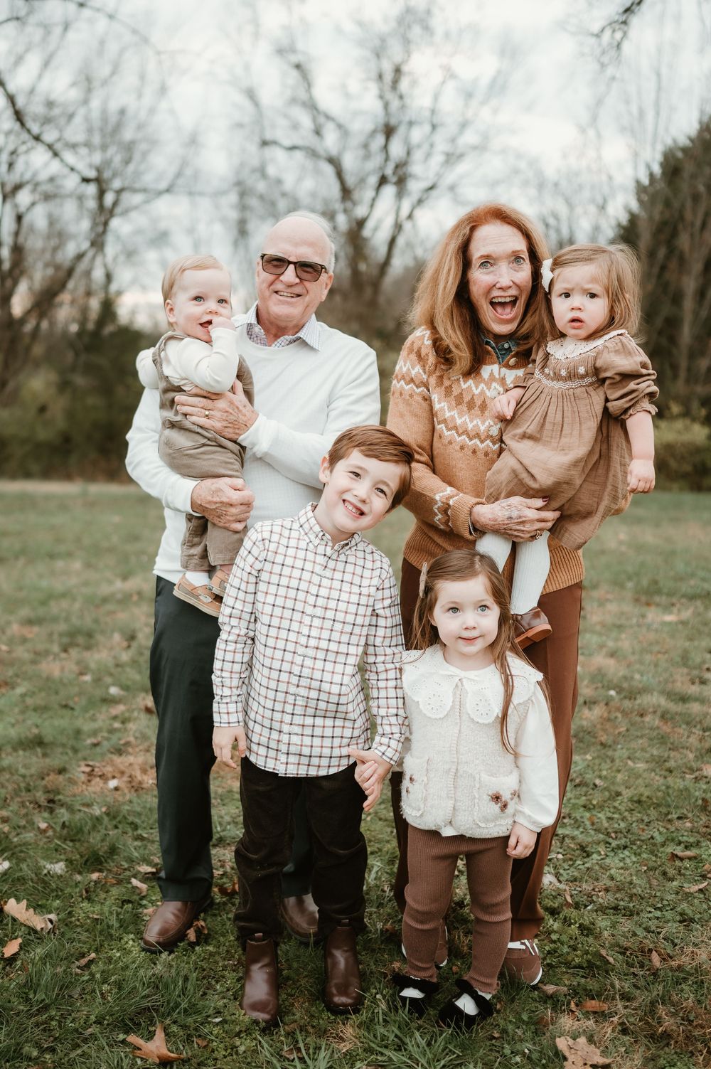 Grandparents smiling outdoors holding toddlers with two young children standing in front on green grass.