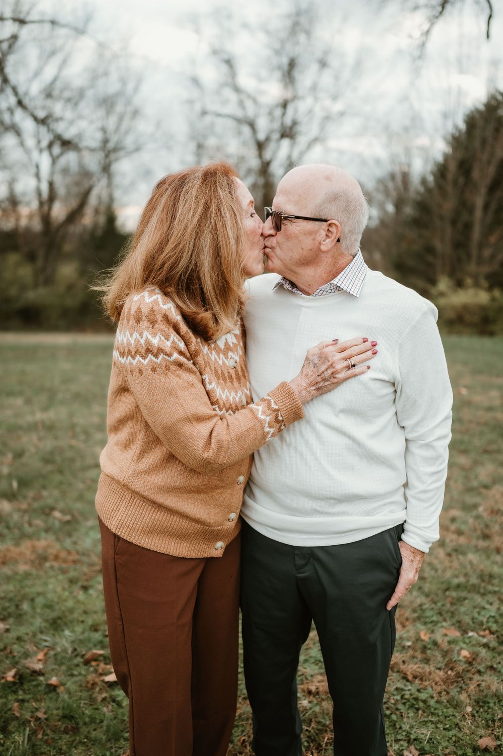An older couple shares a tender kiss outdoors in a park, wearing cozy fall sweaters amid leafless trees.