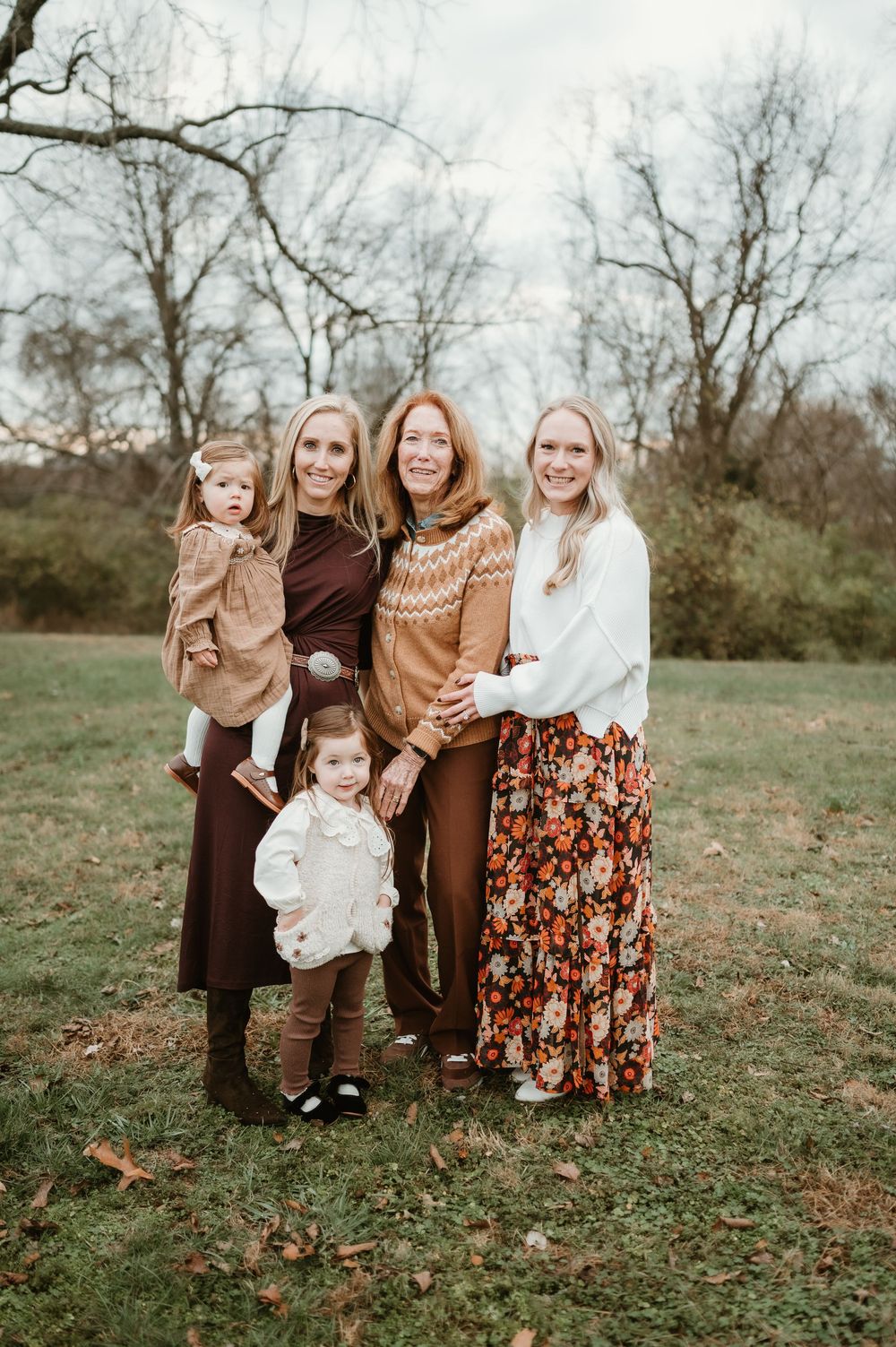 A multigenerational family of women and two young girls pose outdoors in autumn, wearing warm earth-toned outfits.