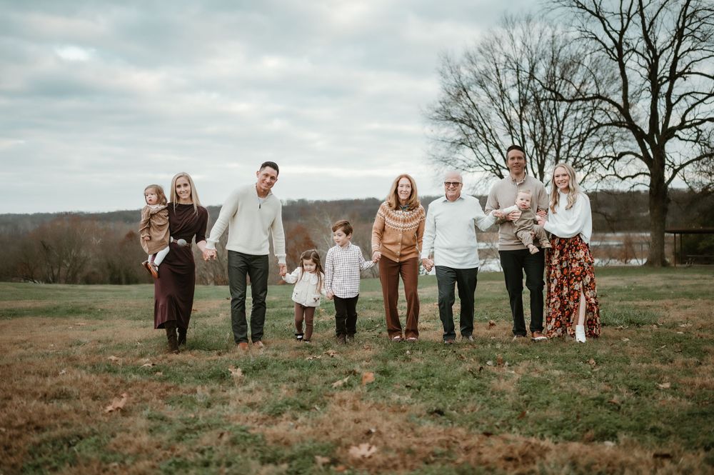 Large family posing outdoors in a field during fall, holding hands and smiling in coordinated earth-tone outfits.
