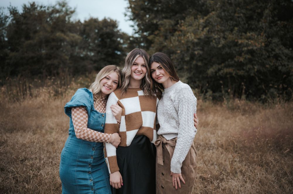 Three friends embrace while wearing coordinated outfits in an autumn outdoor setting with trees in the background.