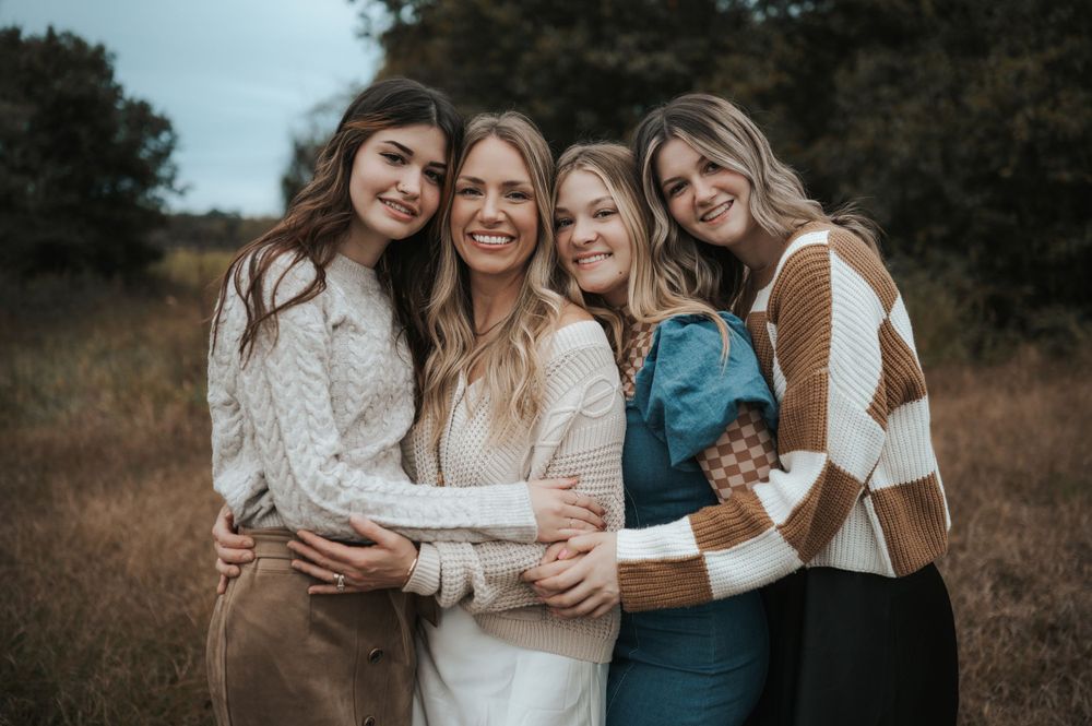 A group of four friends in sweaters embrace while smiling together in a rustic outdoor setting during autumn.