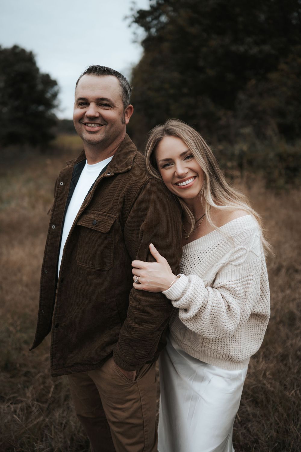 A couple embraces in a rustic outdoor setting wearing brown and white sweaters on a moody autumn day.