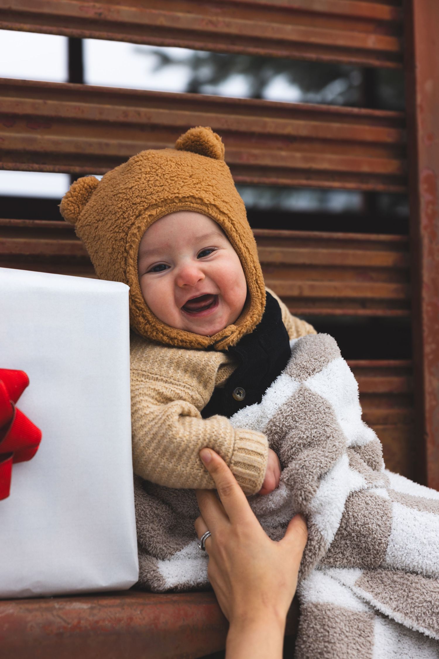 Happy laughing baby wearing a brown bear hat and wrapped in a blanket sits near a Christmas gift.