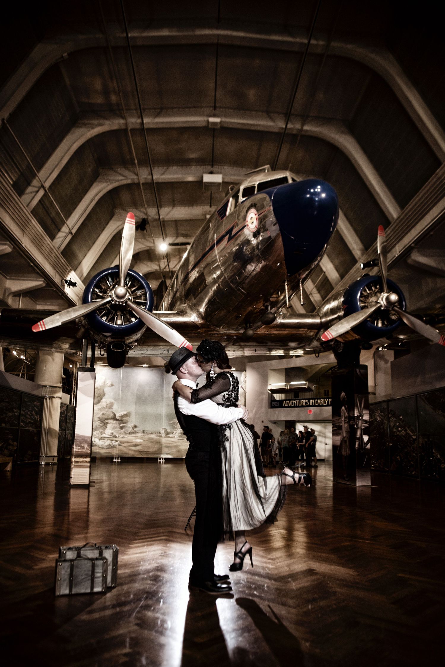 Romantic vintage-style couple embraces in dramatic lighting in front of historic aircraft at an aviation museum.