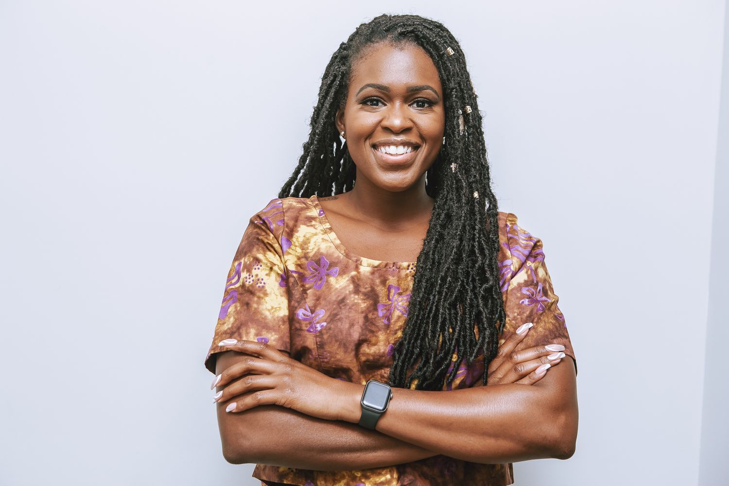 A confident person with long braids wearing a brown floral top poses with arms crossed against a white wall.
