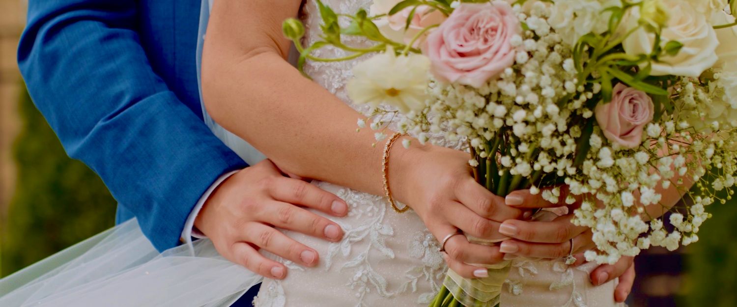 Close-up view of hands holding a delicate pink rose and baby's breath wedding bouquet with white lace dress details.