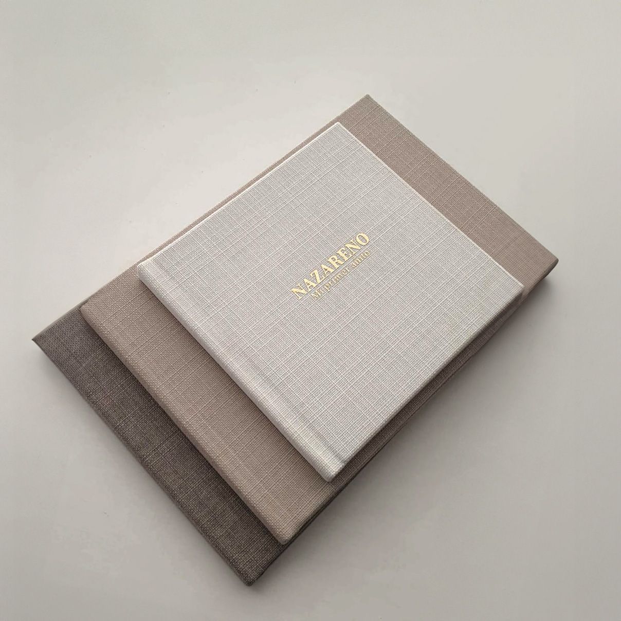 Three elegant cloth-bound notebooks in neutral taupe and gray tones with gold lettering stacked together.