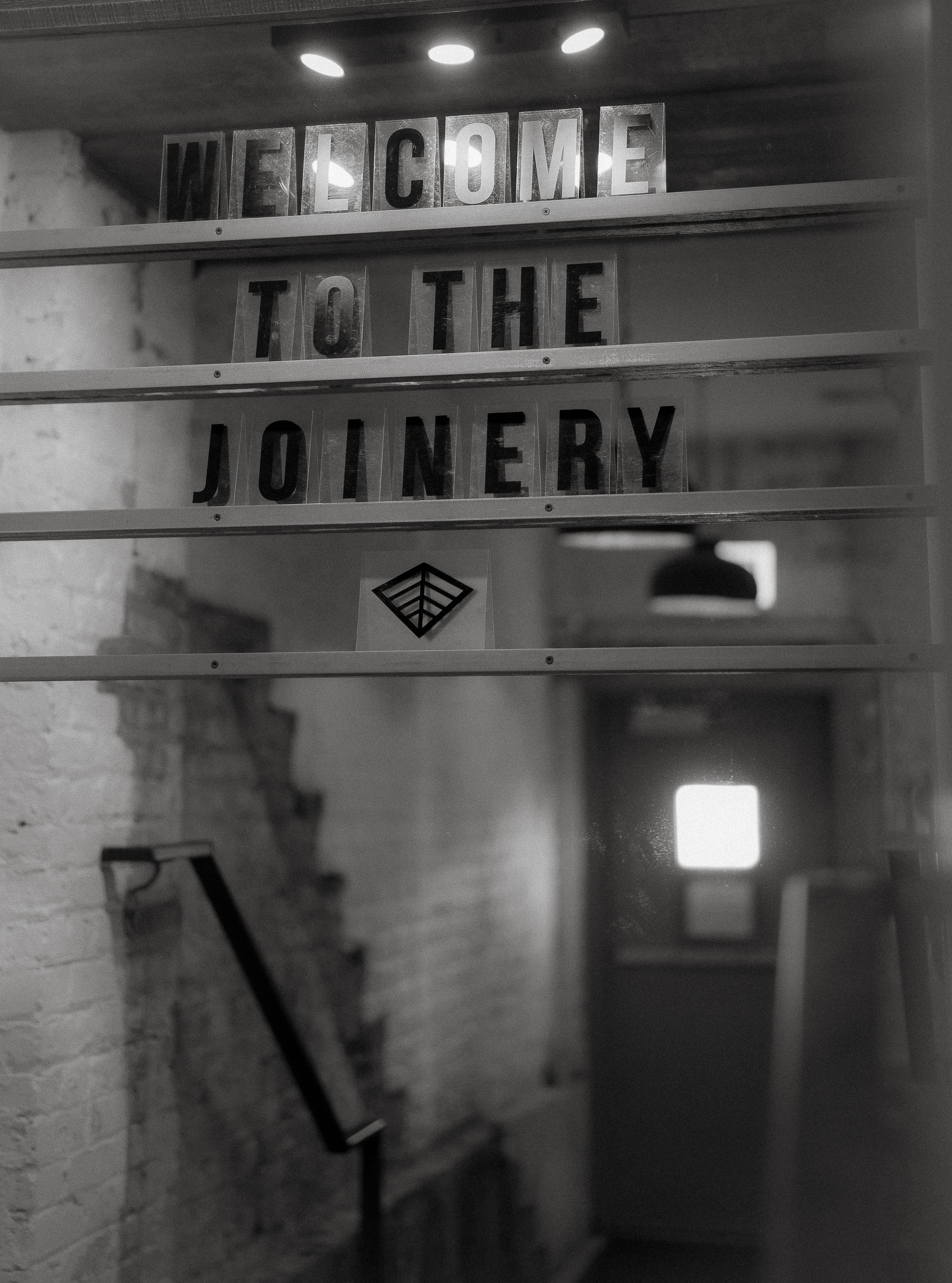 A black and white photo shows illuminated letters reading 'Welcome to the Joinery' above a dimly lit stairway entrance.