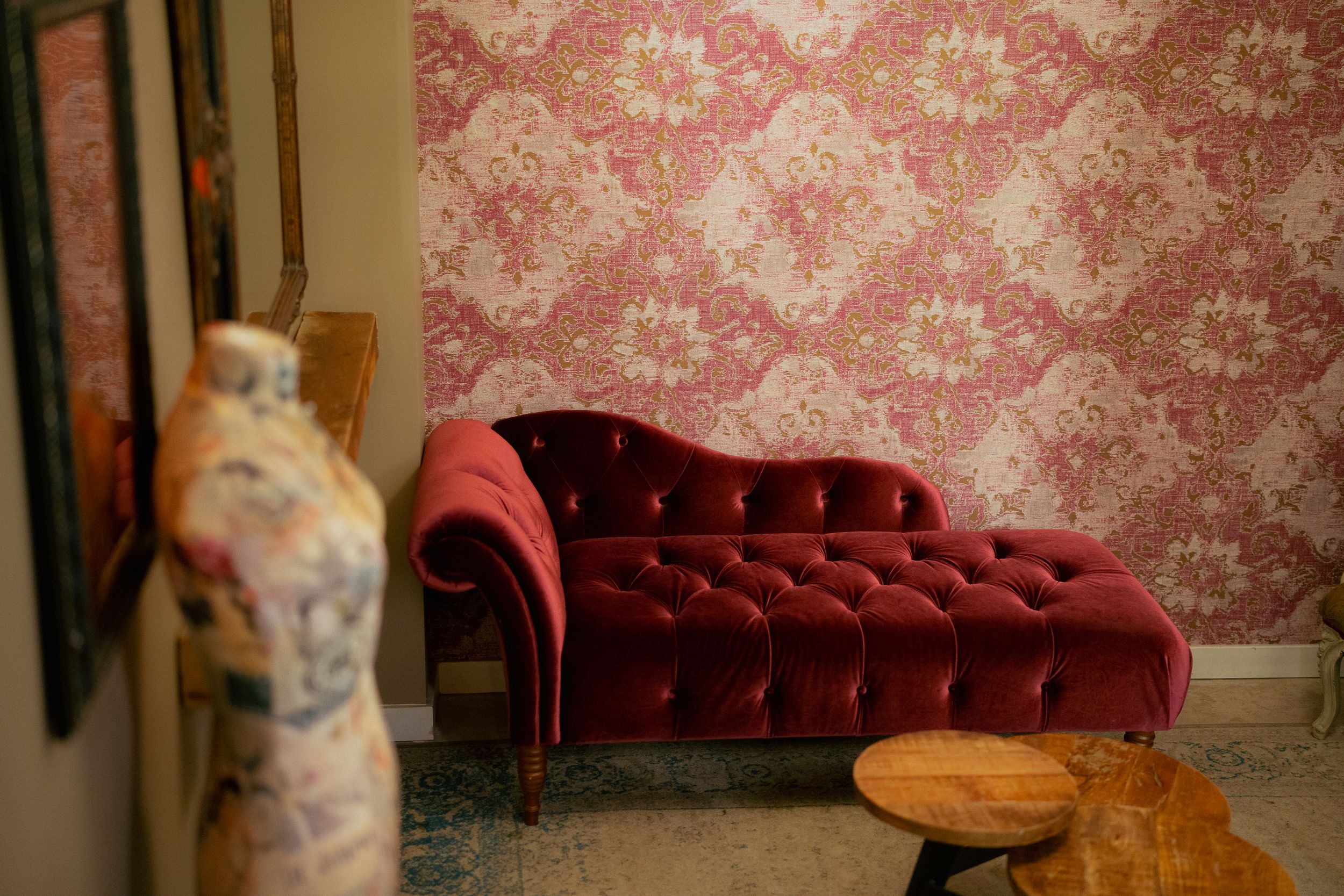 Elegant red velvet chaise lounge against pink damask wallpaper with decorative mannequin and wooden stool.