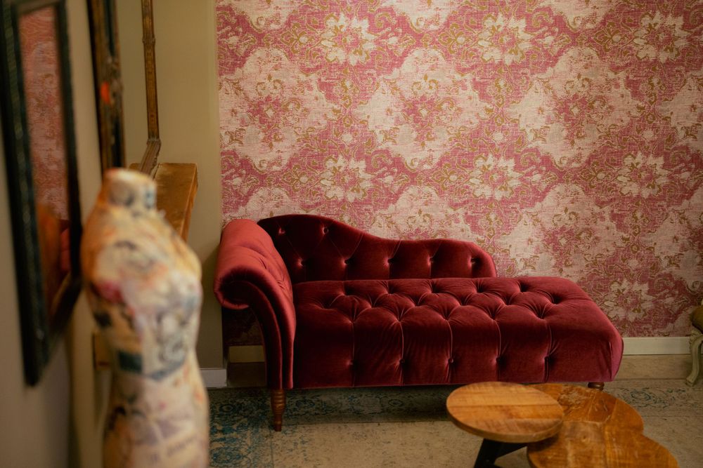 Elegant red velvet chaise lounge against pink damask wallpaper with decorative mannequin and wooden stool.