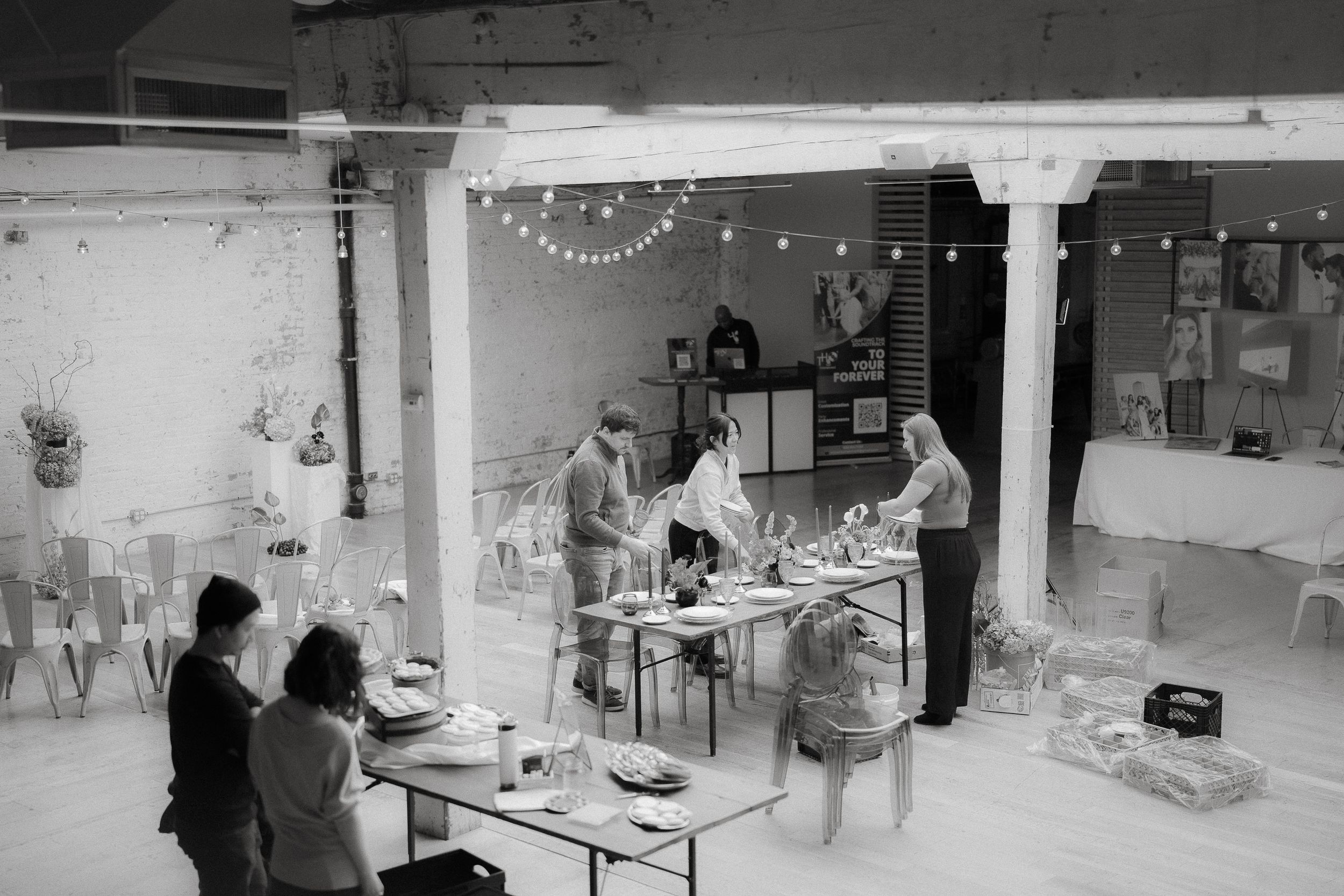A black and white photograph of people working in a studio space with string lights and industrial decor.