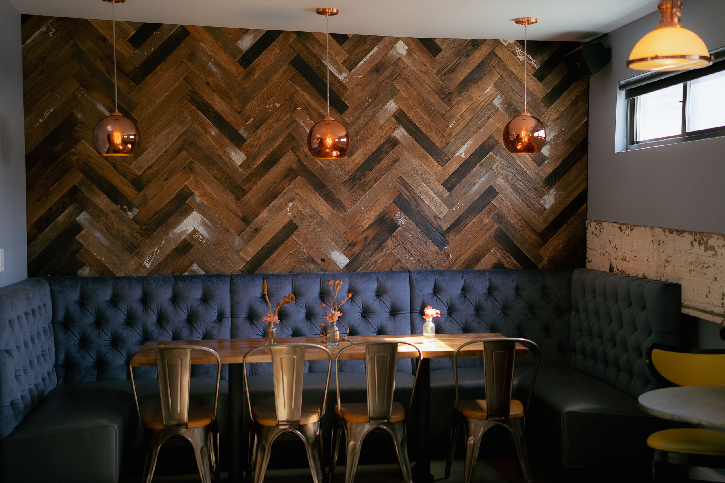 Modern restaurant booth with navy tufted seating, herringbone wood wall panels, and copper pendant lights.