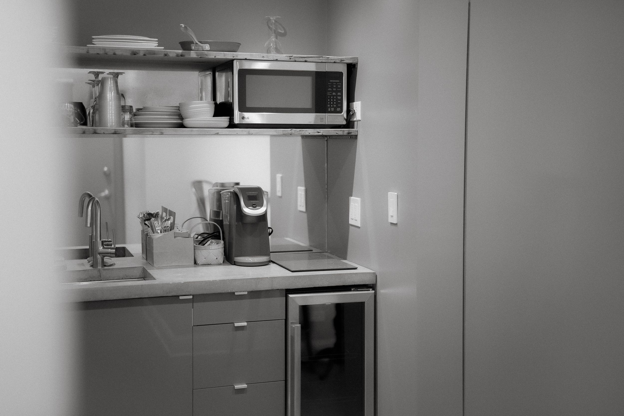 Black and white photograph of a small kitchen area with microwave, coffee maker, and minimalist storage shelves.