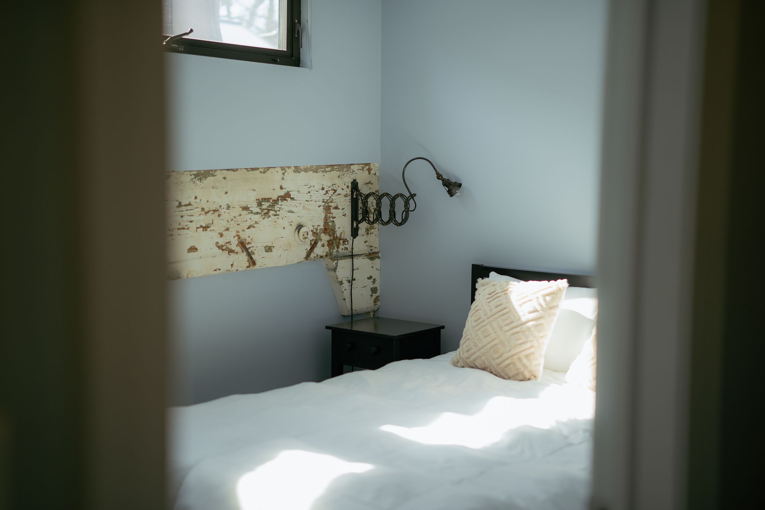 Sunlit bedroom with white bedding, vintage headboard, and wall-mounted reading lamp casting soft shadows.