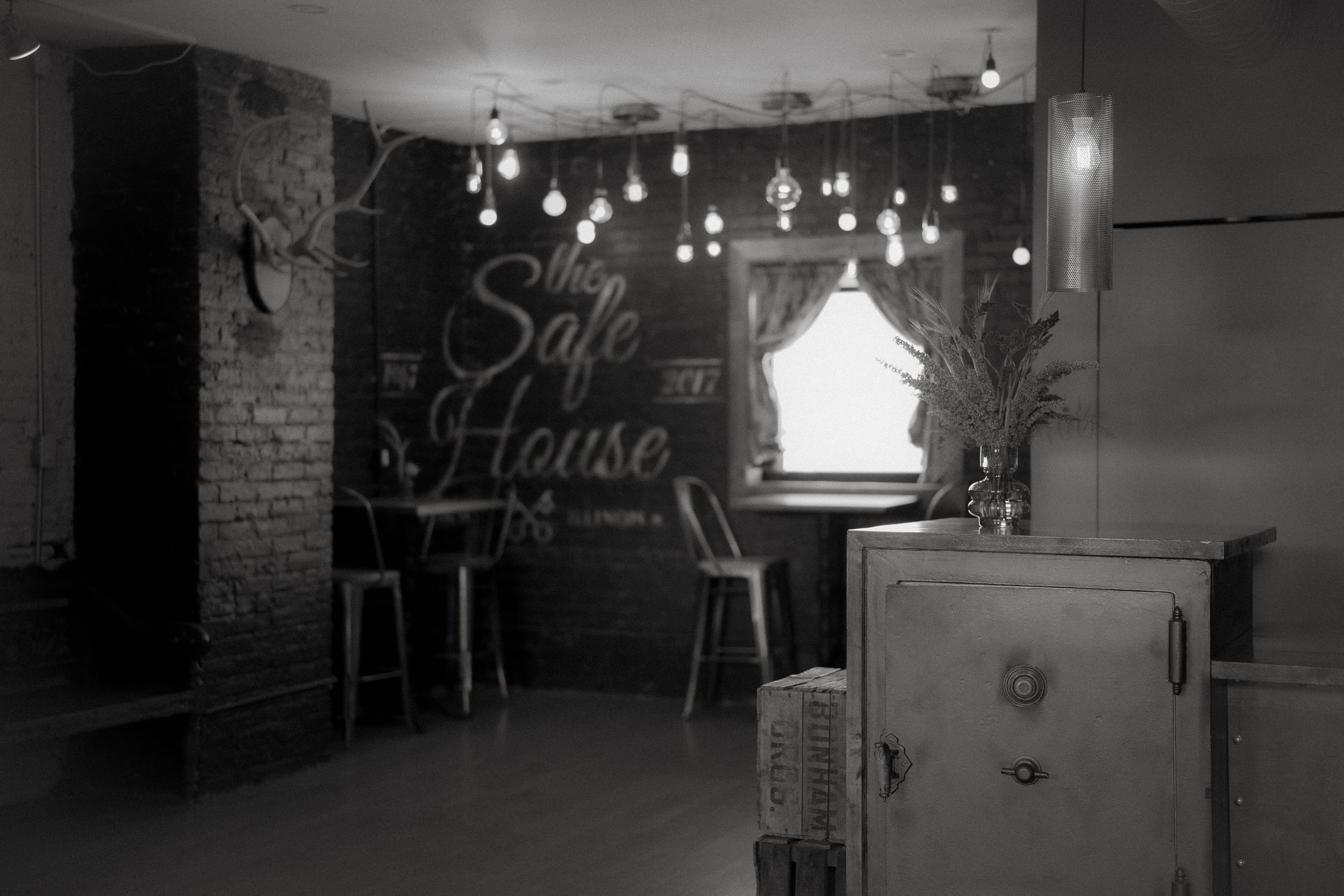 Black and white interior view of a cozy space with hanging lights and a chalkboard wall displaying 'House' text.