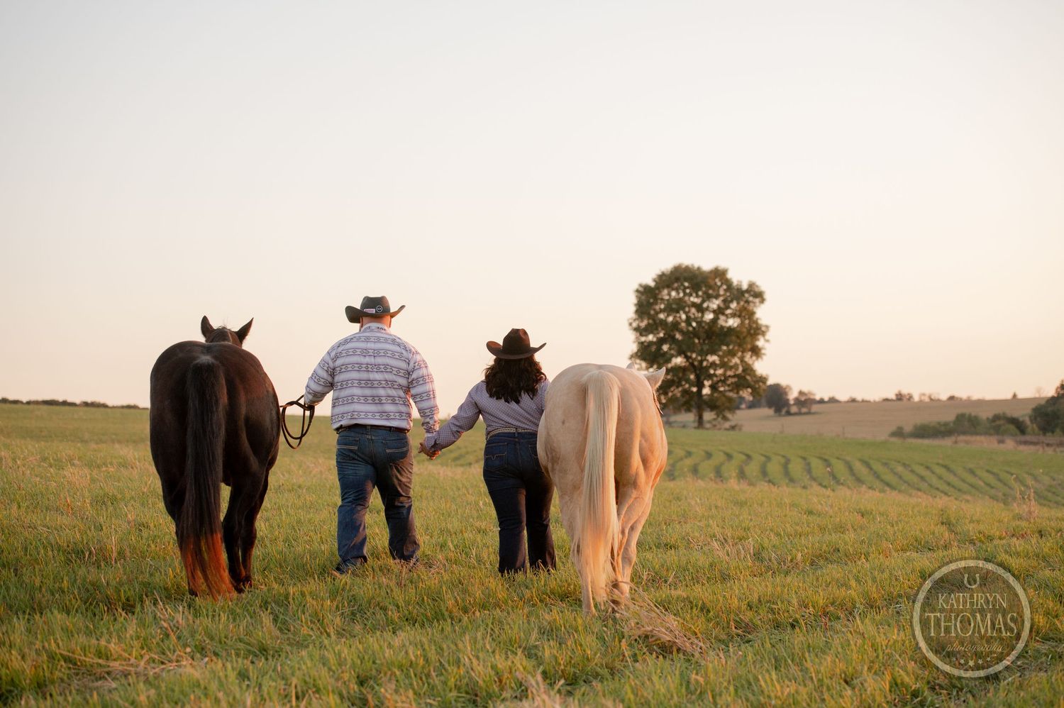 Two people walking with their horses through a scenic countryside field at sunset.