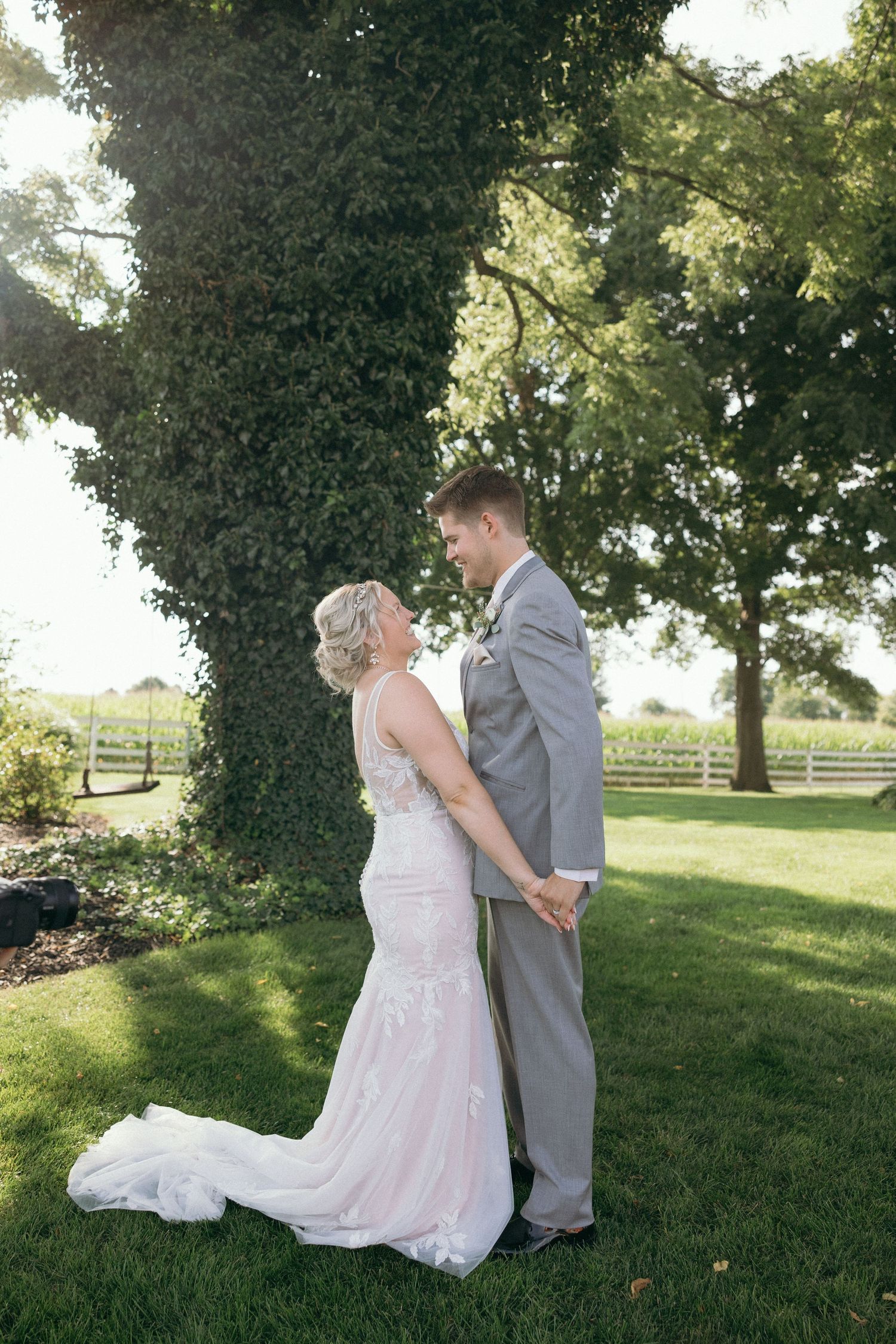 A romantic outdoor wedding moment captured on a sunny day under lush green trees and wooden fences.