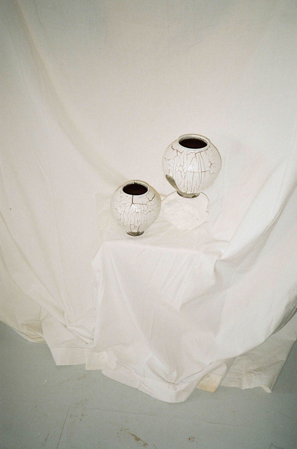 Two white crackle-glazed ceramic vases with dark interiors displayed on white draped fabric backdrop.