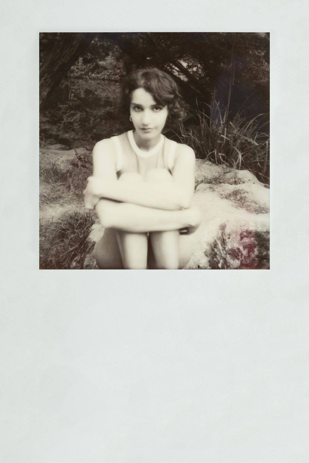 Artistic black and white photograph showing a person sitting outdoors in a contemplative pose against a natural background.