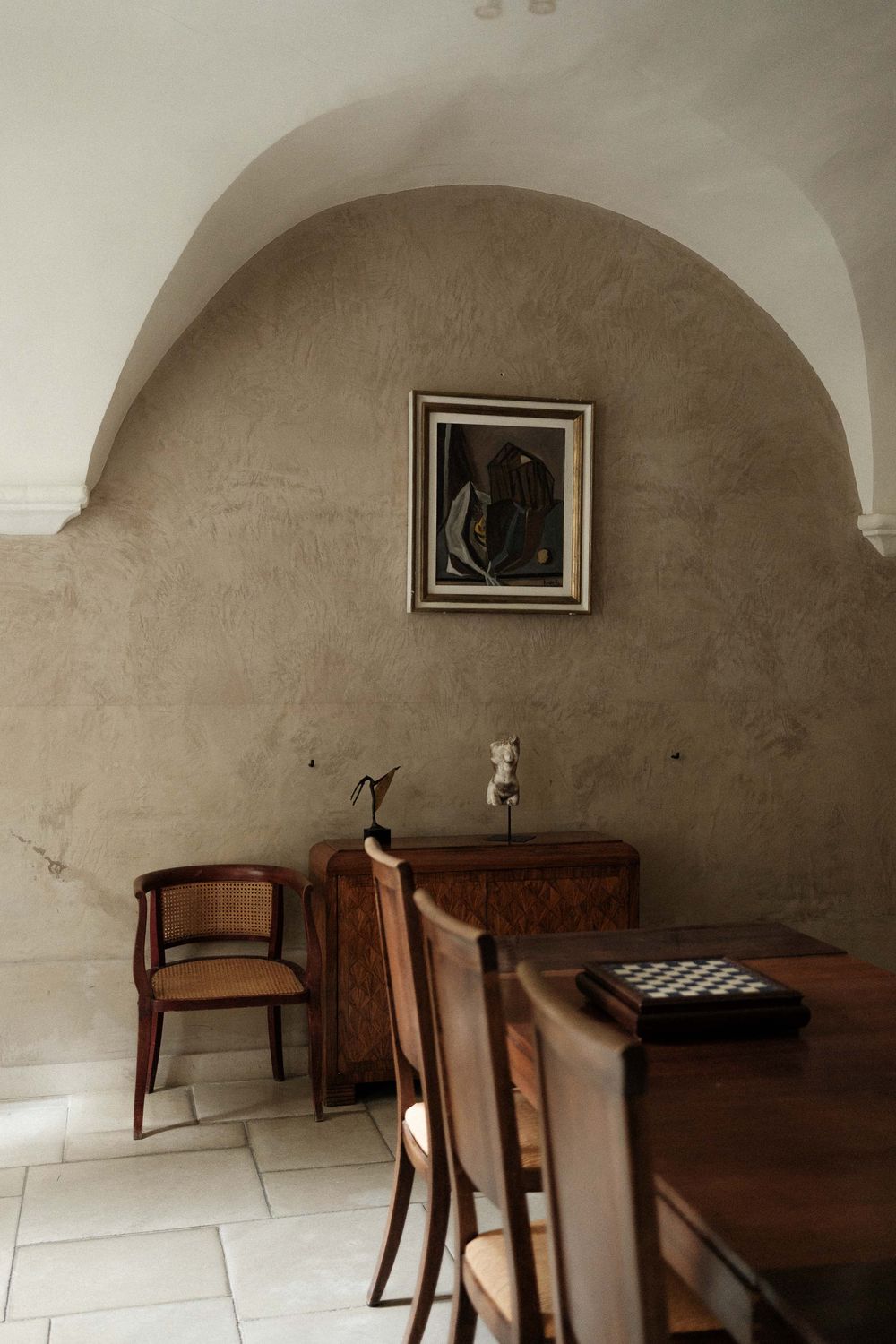 Rustic arched alcove with antique wooden furniture, a framed artwork, and a chess board in an elegant stone-walled room.
