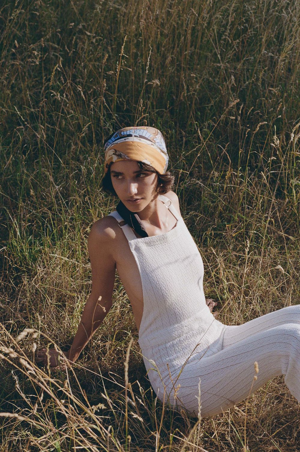 A person in a white summer dress and headband sits gracefully among tall dried grass in golden afternoon light.