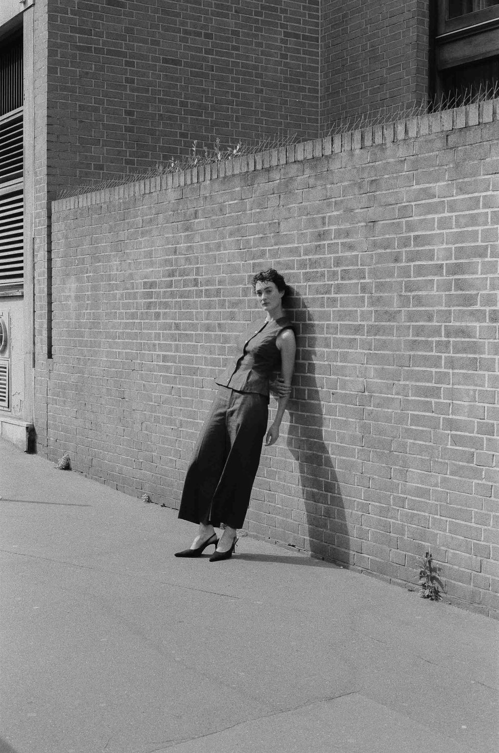 A vintage black and white photograph shows a woman leaning against a brick wall wearing a dress and heels.