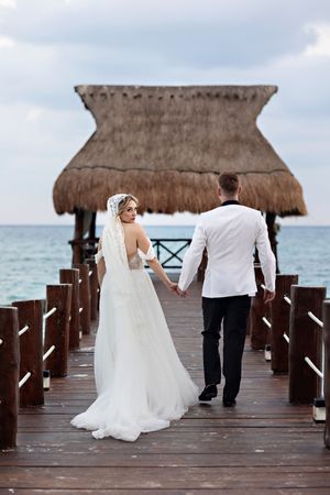A couple walks on a wooden pier with thatched palapa and ocean view at a tropical destination wedding.