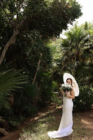 A figure in a white wedding dress holds a bouquet while standing in a lush garden with palm trees and greenery.