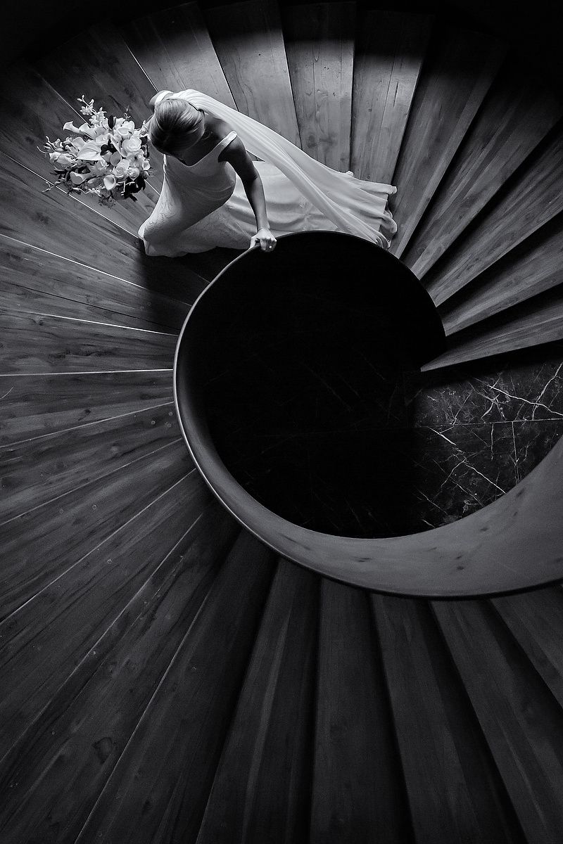 A black and white aerial view of a spiral staircase with flowing white fabric and flowers visible from above.