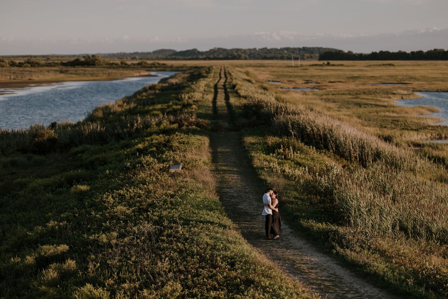 Long shadows stretch across a coastal path as someone walks through tall grass at sunset.