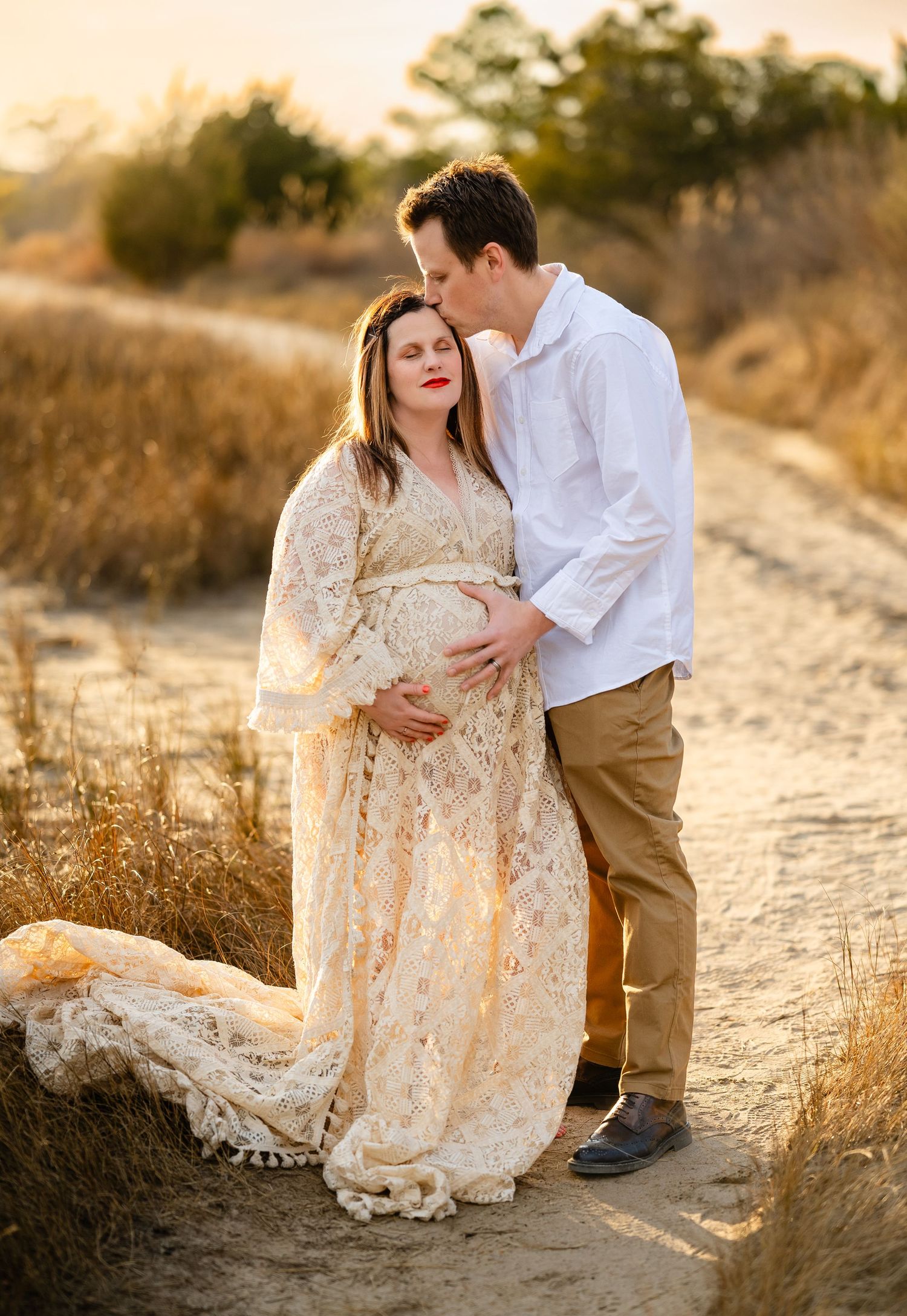 A couple shares an intimate moment on a dirt path at sunset while wearing a lace dress and white shirt.